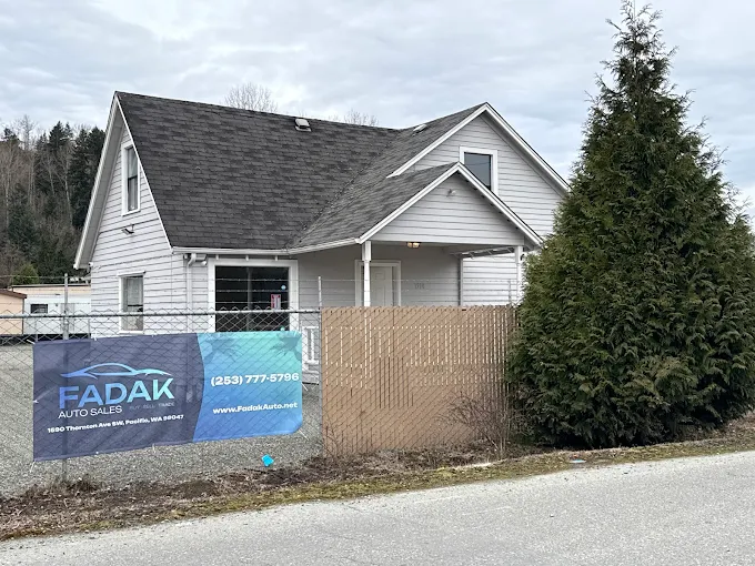 White house with Fadak Auto Sales sign, evergreen tree, chain-link fence