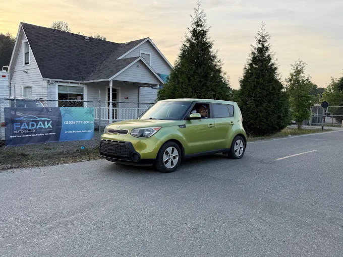 Green Kia Soul parked on street near white house and auto sales sign