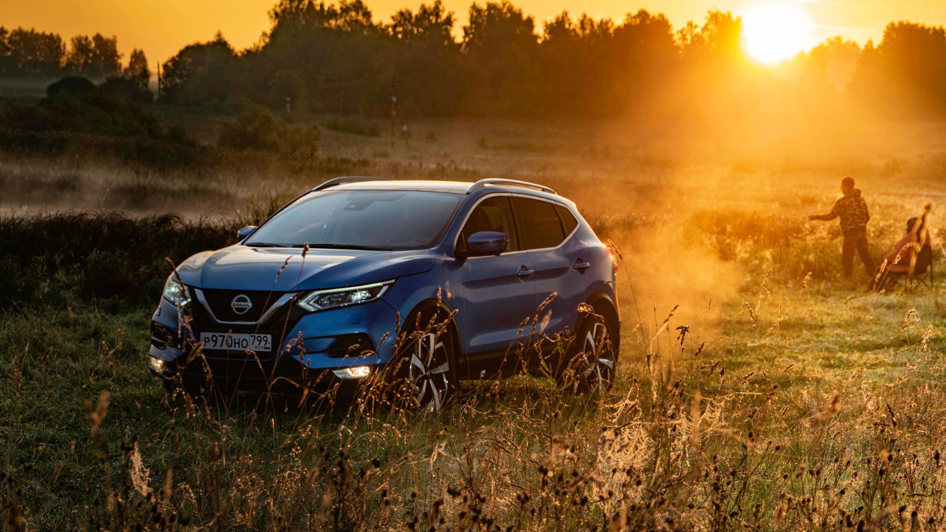 Blue Nissan SUV in grassy field at golden sunset with people in background