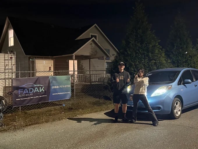 Two people standing by blue electric car at night near Padak Auto Sales sign