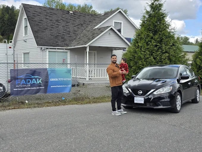 Man holding child next to black Nissan car in front of auto sales business