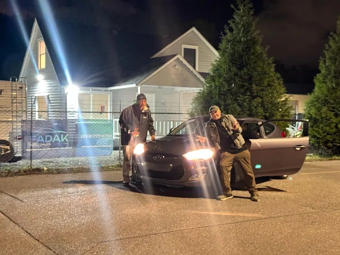 Two people standing by car at night near suburban house with bright lights